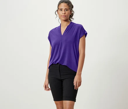 Eine Frau steht und trägt ein lila Blusenshirt mit V-Ausschnitt, schwarze Shorts und weiße Turnschuhe.
