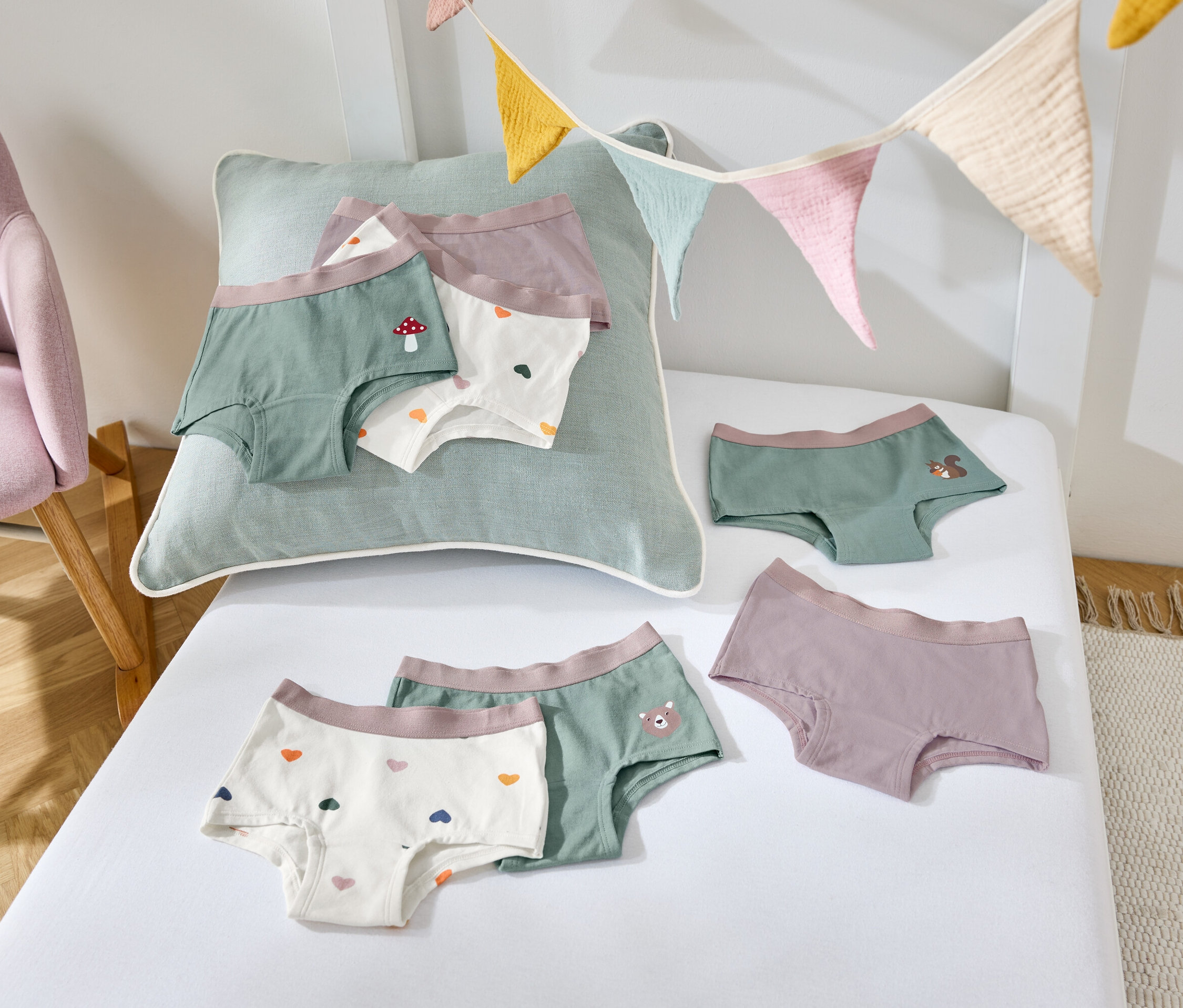 Auf einem weißen Laken liegen Kinderunterhosen in verschiedenen Farben und Mustern, daneben steht ein lila Sessel.