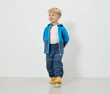 Ein Junge in einer blau-grauen Jacke mit Kapuze, Hose und gelben Gummistiefeln steht vor einer weißen Wand.