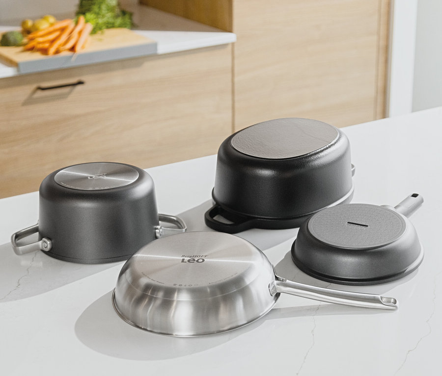 Vier Pfannen liegen mit dem Boden nach oben auf einer weißen Küchentheke. Im Hintergrund sind eine hölzerne Küchenzeile und Gemüse auf einem Schneidebrett zu sehen.