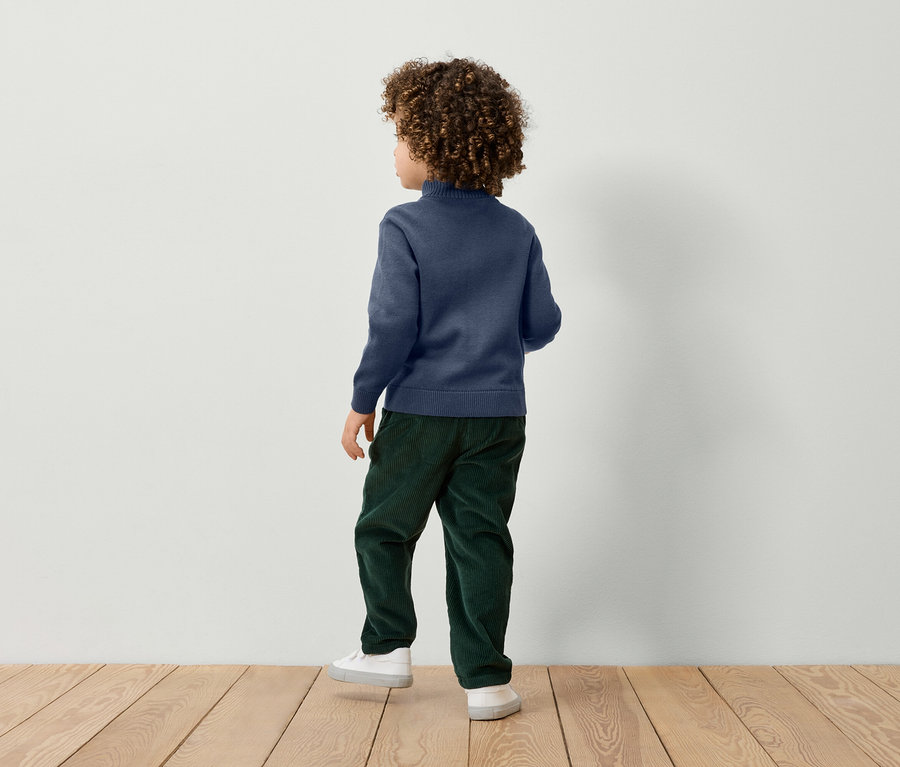 Junge mit lockigem Haar steht mit dem Rücken zum Betrachter und trägt einen blauen Pullover, eine grüne Hose und weiße Turnschuhe.