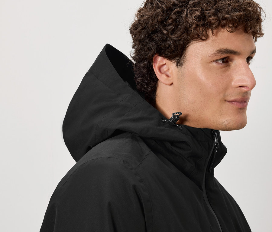 Ein Mann mit braun gelocktem Haar trägt eine schwarze Kapuzenjacke.