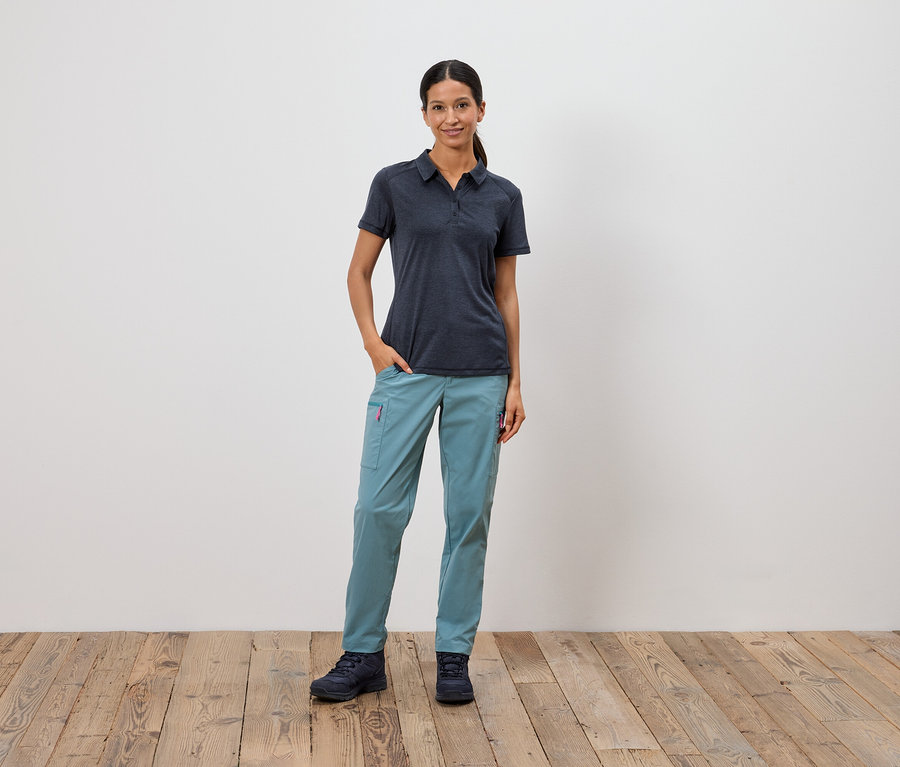 Eine Frau steht mit einer Hand in der Tasche vor einer weißen Wand.