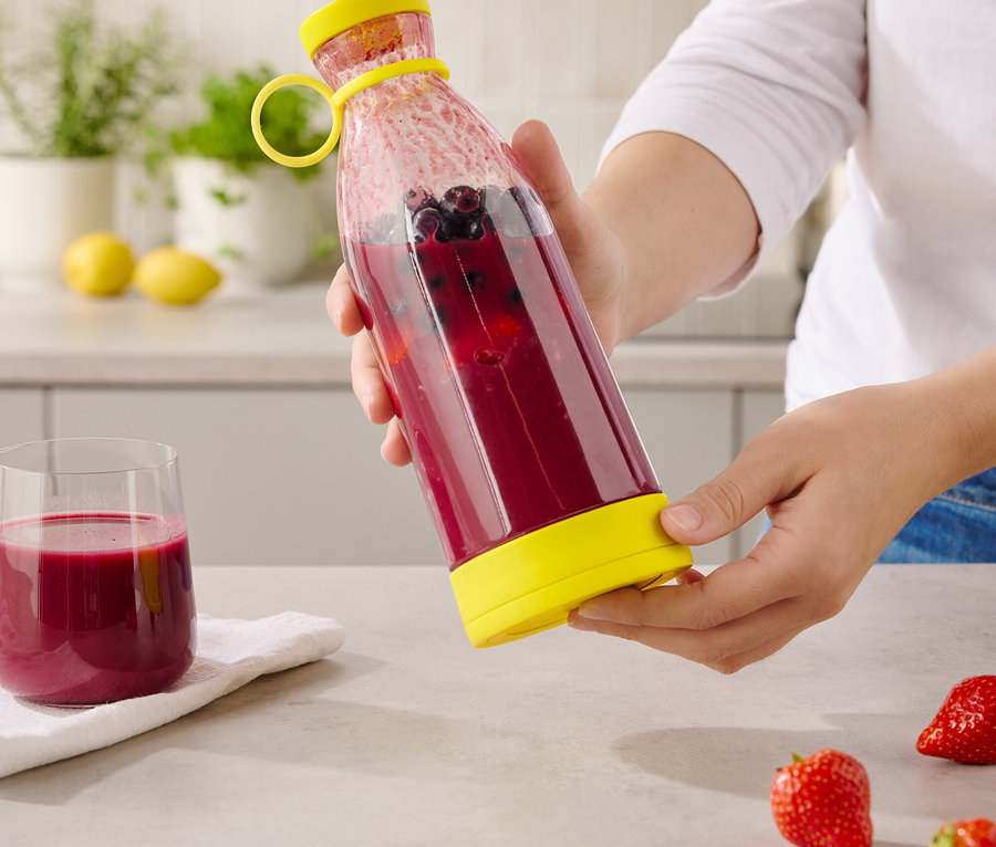 Eine Person hält eine durchsichtige Flasche mit einem gelben Deckel und Boden, gefüllt mit einem roten Getränk und Blaubeeren.