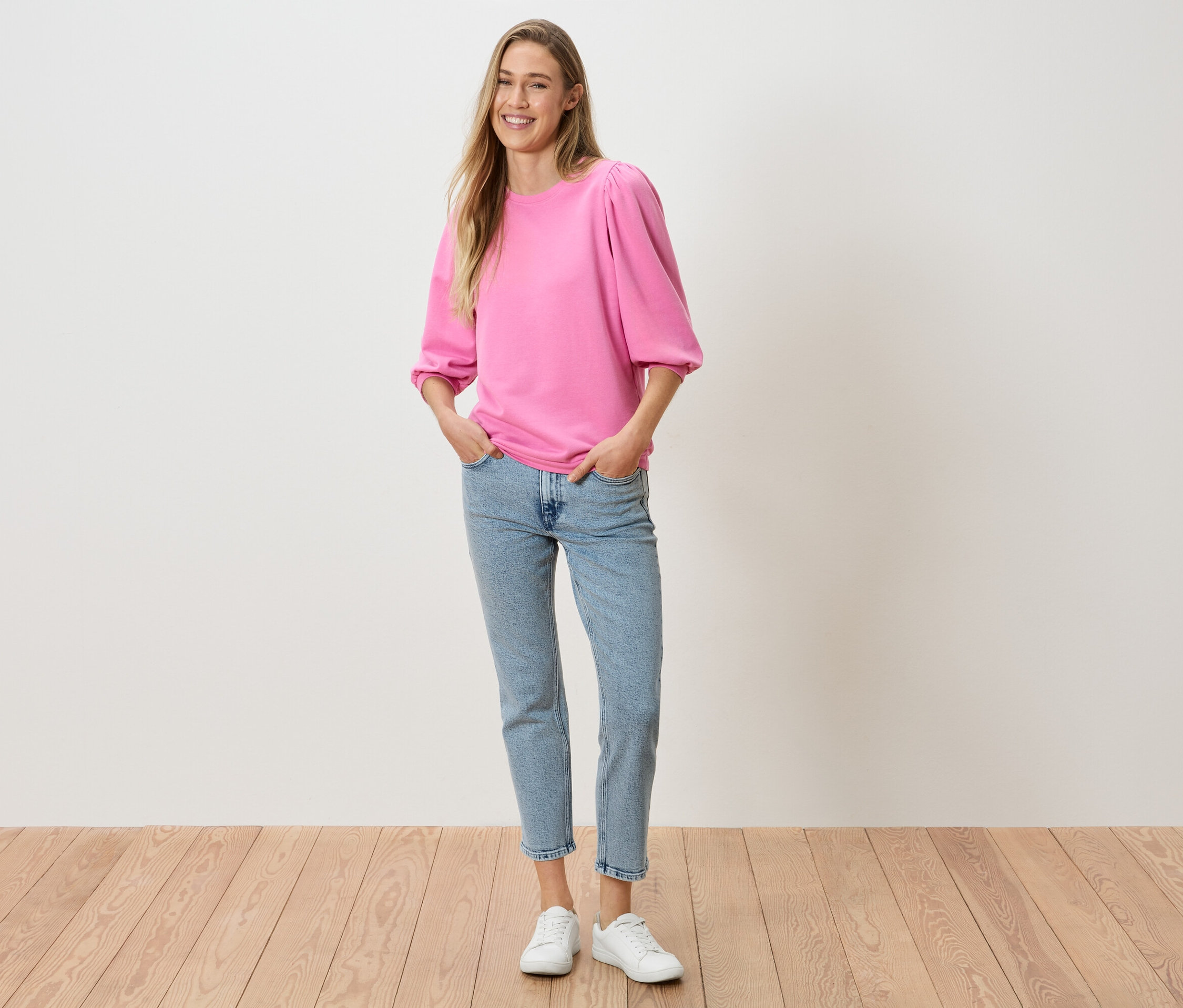 Frau mit ihren Händen in den Hosentaschen, trägt ein pinkes Oberteil mit Puffärmeln, Jeans und weiße Turnschuhe.