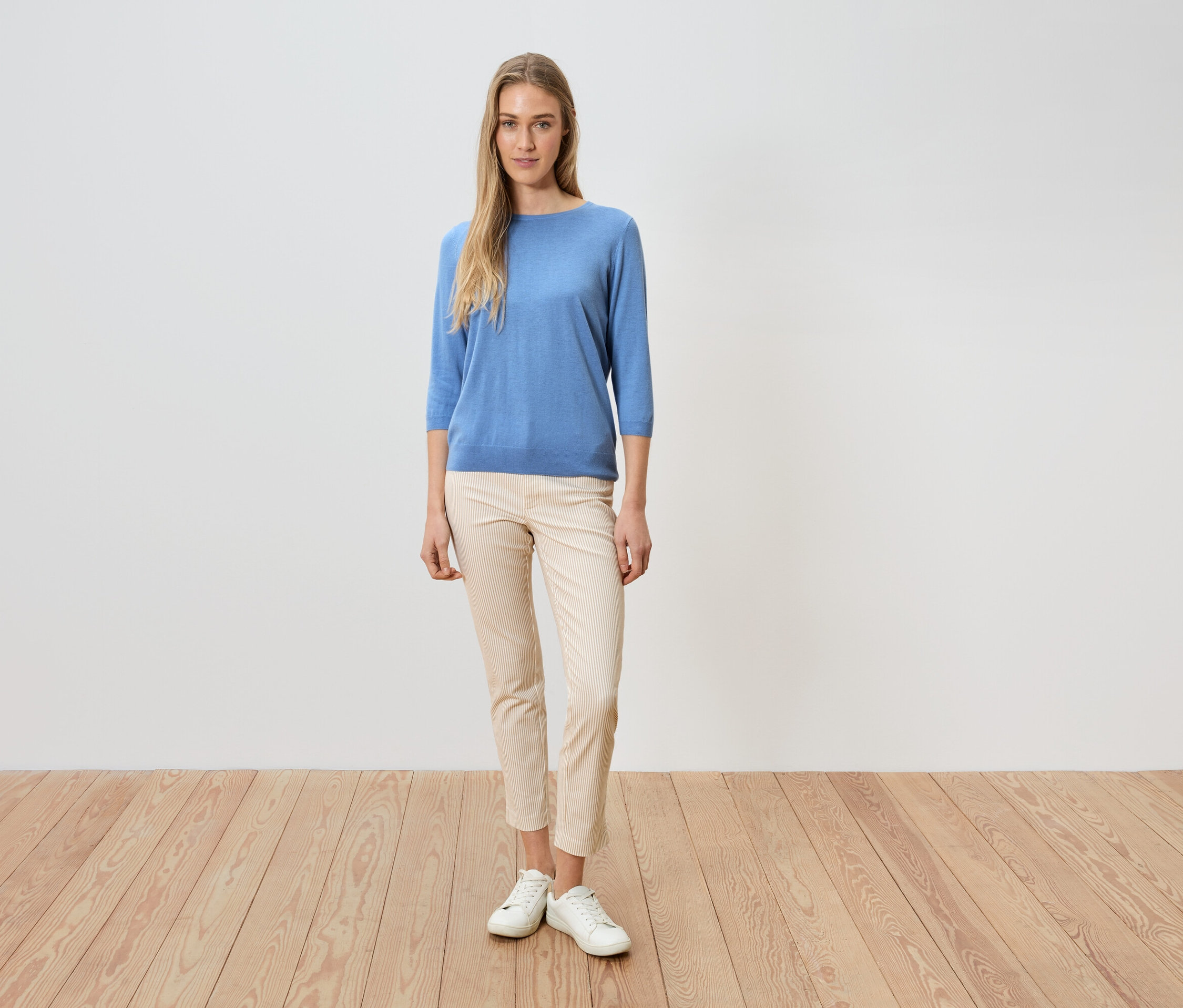 Eine Frau steht vor einer weißen Wand, sie trägt einen blauen Pullover, eine helle Hose und weiße Schuhe.