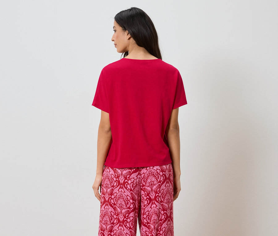 Rückansicht einer Frau mit dunklen Haaren, die ein rotes T-Shirt und rosafarbene, gemusterte Hosen trägt.