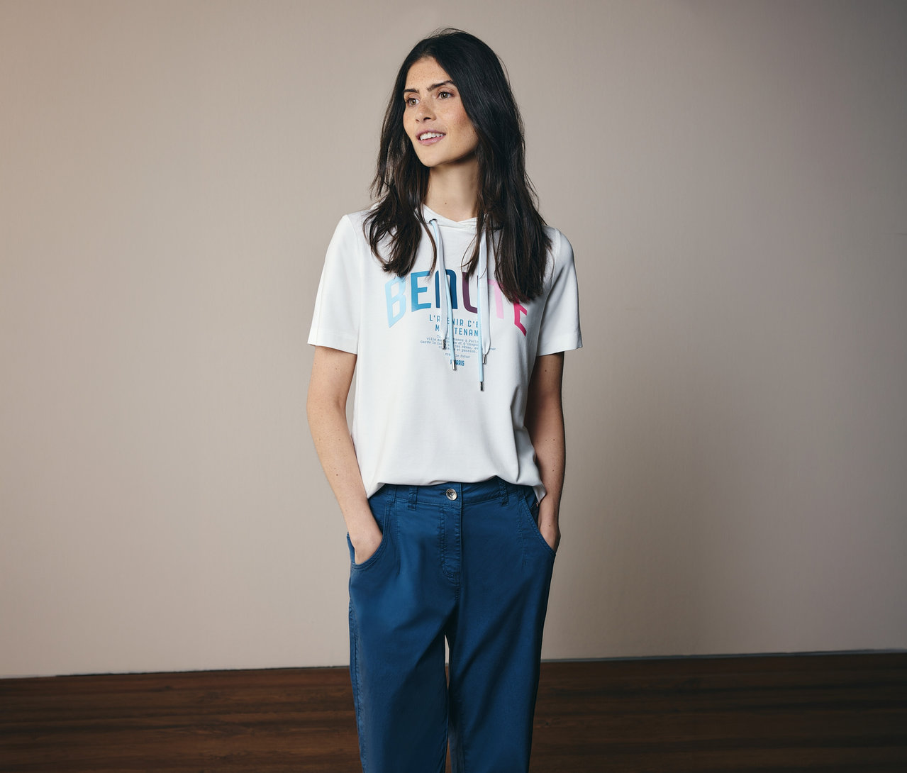Frau mit blauen Hosen und weißem CECIL Hoodie-Shirt mit Aufdruck und Kapuze, die Hände in den Hosentaschen.