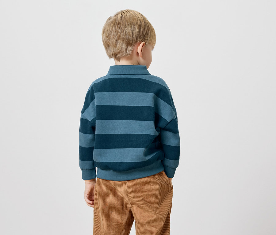 Rückansicht eines Kindes mit blonden Haaren, das ein blau gestreiftes Langarmshirt und eine braune Cordhose trägt.