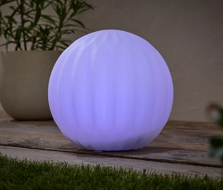 Eine runde, lila Lampe steht auf einem Holztisch, neben einer Topfpflanze.