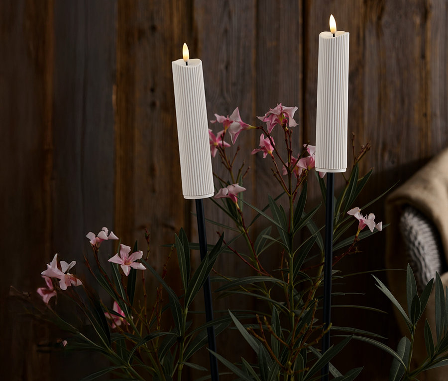 Zwei weiße, gerippte Kerzen auf schwarzen Ständern inmitten von rosa Blüten und grünen Blättern vor einem dunklen Holzhintergrund.