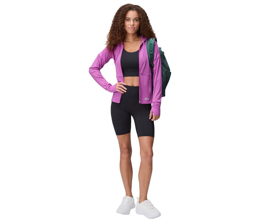 Frau mit lockigem Haar, schwarzem Sport-BH, schwarzen Radlershorts, lila Fila-Jacke und weißen Turnschuhen posiert mit grüner Tasche.
