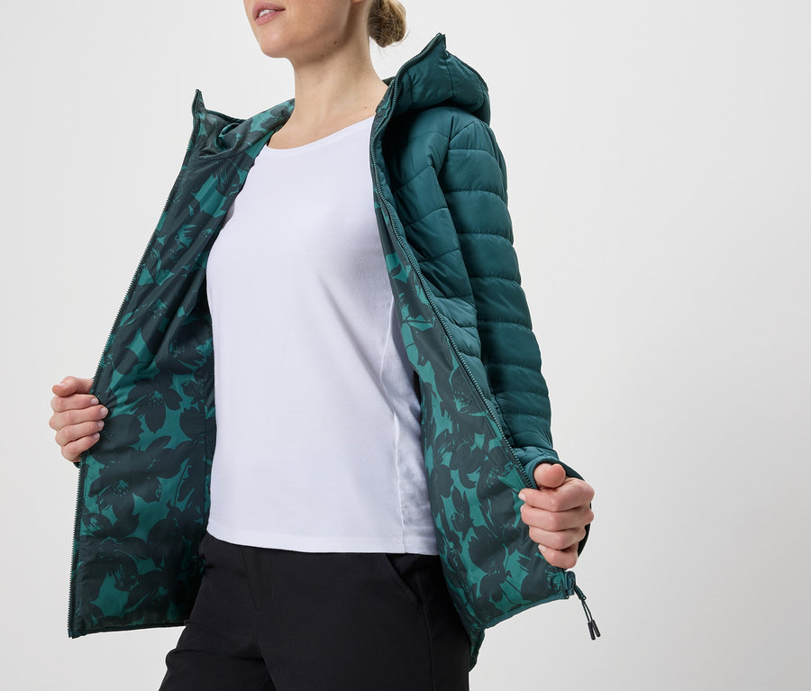 Frau mit geöffneter, petrolfarbener Steppjacke mit Kapuze und Blumenmuster-Futter über einem weißen T-Shirt und schwarzer Hose.