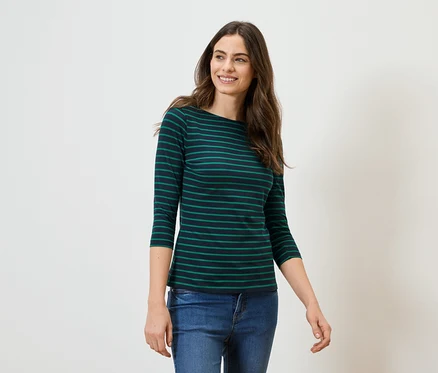 Frau mit langen braunen Haaren trägt ein grün-schwarz gestreiftes Langarmshirt und Jeans vor einem weißen Hintergrund.