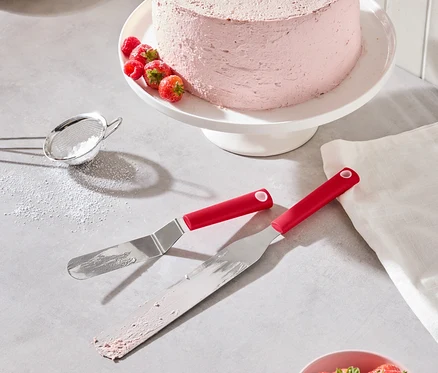 Ein rosa Kuchen mit Erdbeeren und Himbeeren steht auf einem weißen Tortenständer, zusammen mit zwei roten Spateln, einer Schale Erdbeeren und einem Puderzuckersieb.