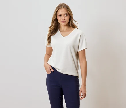 Frau mit langem, gewelltem Haar, trägt ein lockeres, kurzärmeliges, cremefarbenes T-Shirt mit V-Ausschnitt und marineblaue Hosen, eine Hand in der Hosentasche.