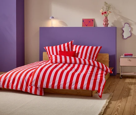 Bett mit rot-weiß gestreifter Bettwäsche und mehreren Kissen vor lila Wand.