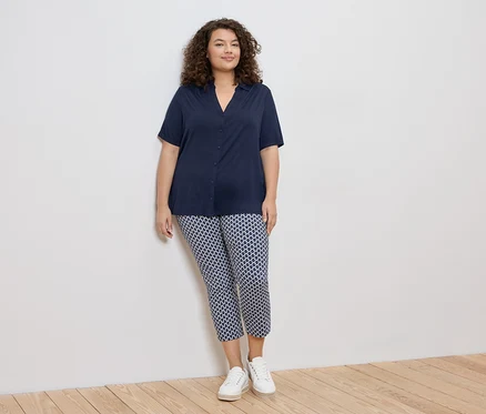 Frau mit lockigem Haar, die eine marineblaue Bluse und gemusterte Hosen mit weißen Turnschuhen trägt und an einer weißen Wand lehnt.