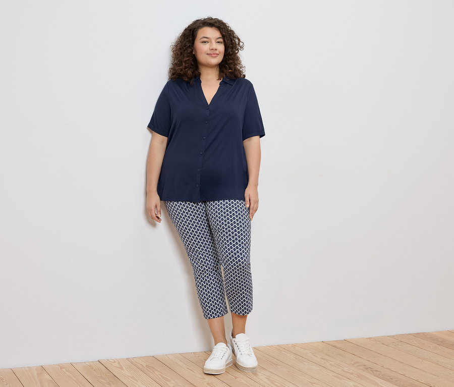 Frau mit lockigem Haar, die eine marineblaue Bluse und gemusterte Hosen mit weißen Turnschuhen trägt und an einer weißen Wand lehnt.
