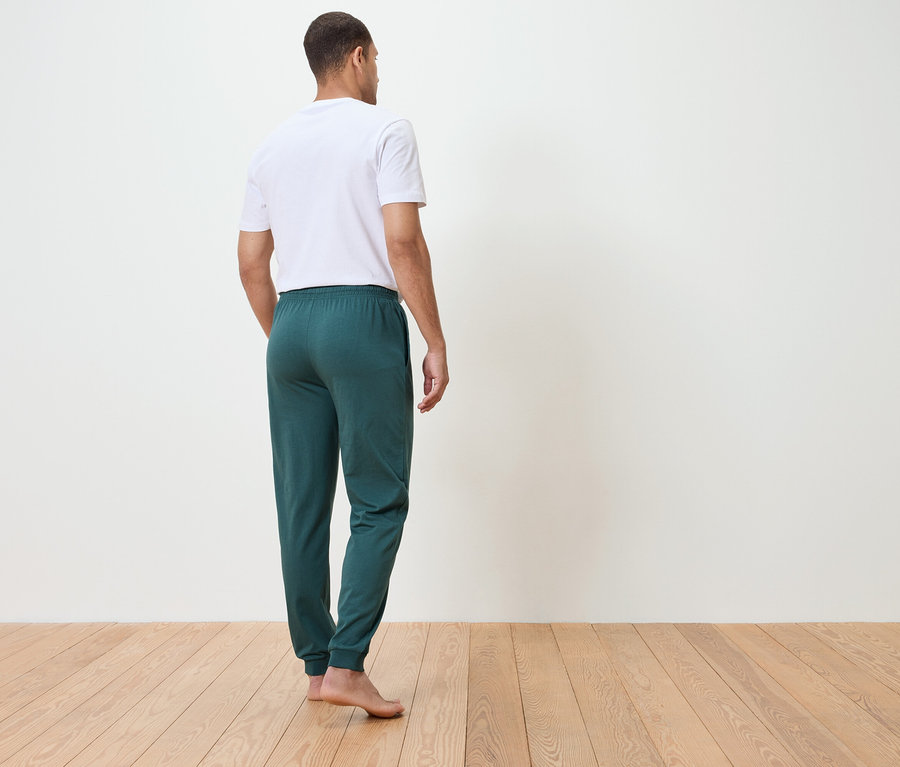 Mann von hinten in einem weißen T-Shirt und grünen Jogginghosen, stehend auf einem Holzboden vor einer weißen Wand.