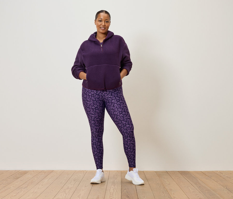 Frau in lila Fleece-Pullover mit halbem Reißverschluss und lila Leggings mit Leopardenmuster, die weiße Turnschuhe trägt.