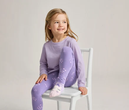 Ein junges Mädchen mit blonden Haaren sitzt auf einem weißen Stuhl, lächelt und blickt nach rechts. Sie trägt ein hellviolettes Langarmshirt, violette Leggings mit hellviolettem Muster und hellrosa Socken mit einem Gesichtsdesign.