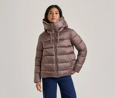 Frau in einer braunen Steppjacke mit hohem Kragen und Kapuze sowie blauen Jeans, die mit den Händen in den Taschen steht.