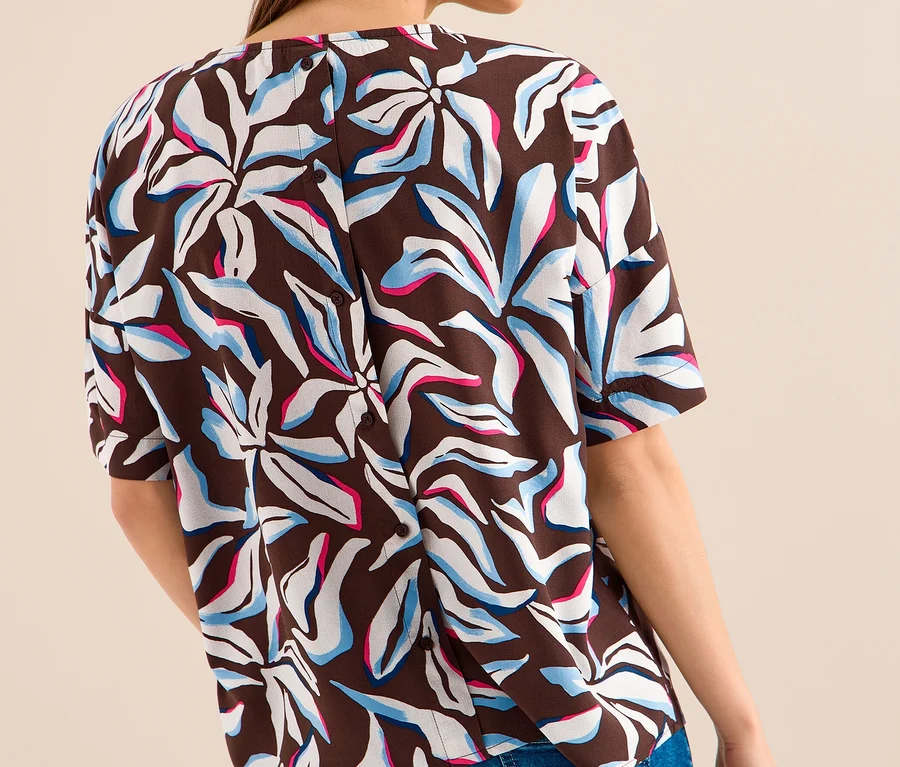 Frau von hinten, in einer braunen CECIL Kurzarmbluse mit weißen, hellblauen und pinken abstrakten Blumenmustern und Knöpfen am Rücken, dazu blaue Jeans.