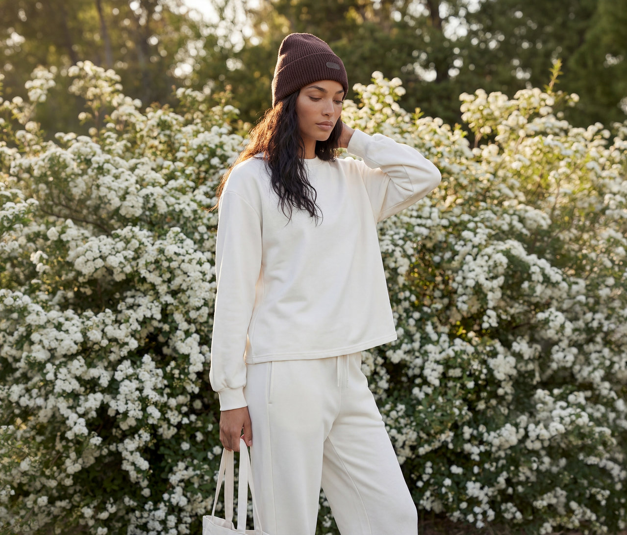 Frau in dunkelbrauner Mütze und cremefarbenem Sweatshirt und Jogginghose, eine Tragetasche haltend, vor einem Busch mit weißen Blüten stehend.
