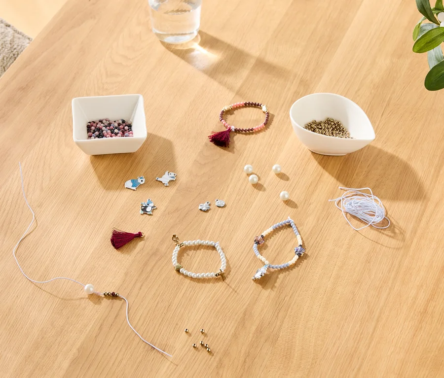 Bastelzubehör zum Herstellen von Armbändern, darunter Perlen in Schalen, lose Perlen, Tieranhänger, Quasten, Faden und fertige/halbfertige Armbänder auf einem Holztisch.