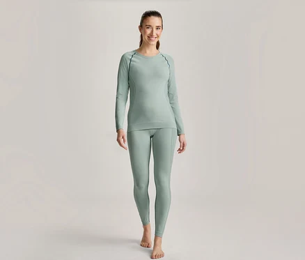 Lächelnde Frau steht in hellgrünem Langarmshirt und passender Leggings mit dunklen Nähten, barfuß.