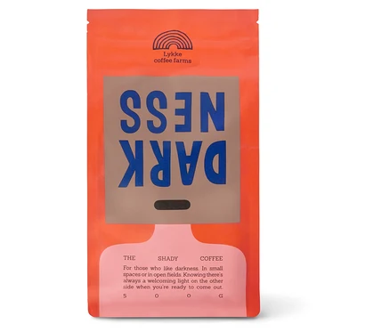 Eine orangefarbene Kaffeepackung mit einem braunen Etikett, auf dem das Wort „DARKNESS“ in blauen Buchstaben steht, wobei „DARK“ auf dem Kopf steht. Darüber befindet sich ein Regenbogenlogo und „Lykke coffee farms“. Darunter steht „THE SHADY COFFEE“ und eine Beschreibung, die mit „500 G“ endet.