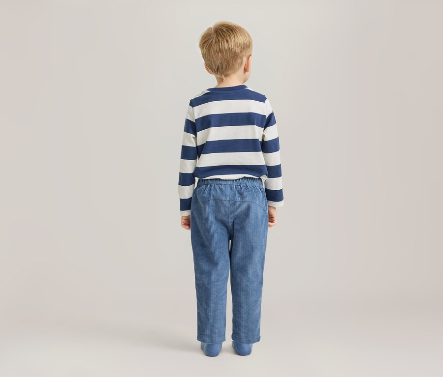 Ein kleiner Junge mit blonden Haaren steht mit dem Rücken zum Betrachter und trägt ein blau-weiß gestreiftes Langarmshirt und blaue Cordhosen.