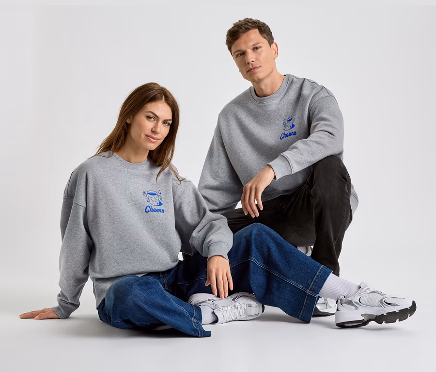 Eine Frau und ein Mann sitzen auf dem Boden und tragen graue Sweatshirts mit der blauen Aufschrift "Cheers".