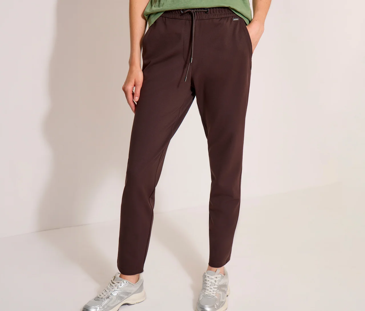 Person trägt eine braune CECIL Jerseyhose, ein grünes T-Shirt und silberne Sneaker, eine Hand steckt in der Hosentasche.