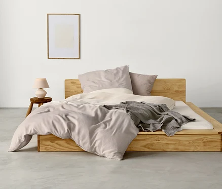 Ein Bett mit hellem Holzrahmen ist mit taupe-offwhite Flanell-Bettwäsche, zwei Kissen und einer grauen Decke bezogen. Links steht ein kleiner Holzhocker mit einer hellrosa Lampe, darüber hängt ein gerahmtes Bild an der weißen Wand.