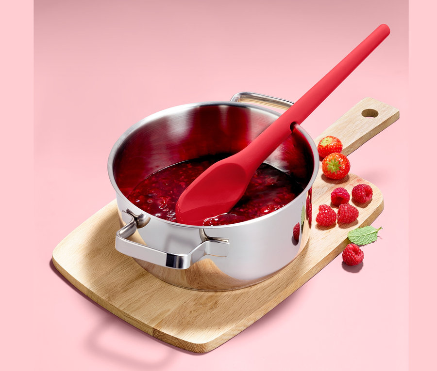 Ein Funktions-Kochlöffel liegt in einem Edelstahltopf mit Marmelade, der auf einem Holzbrett steht. Um ihn herum liegen Himbeeren.