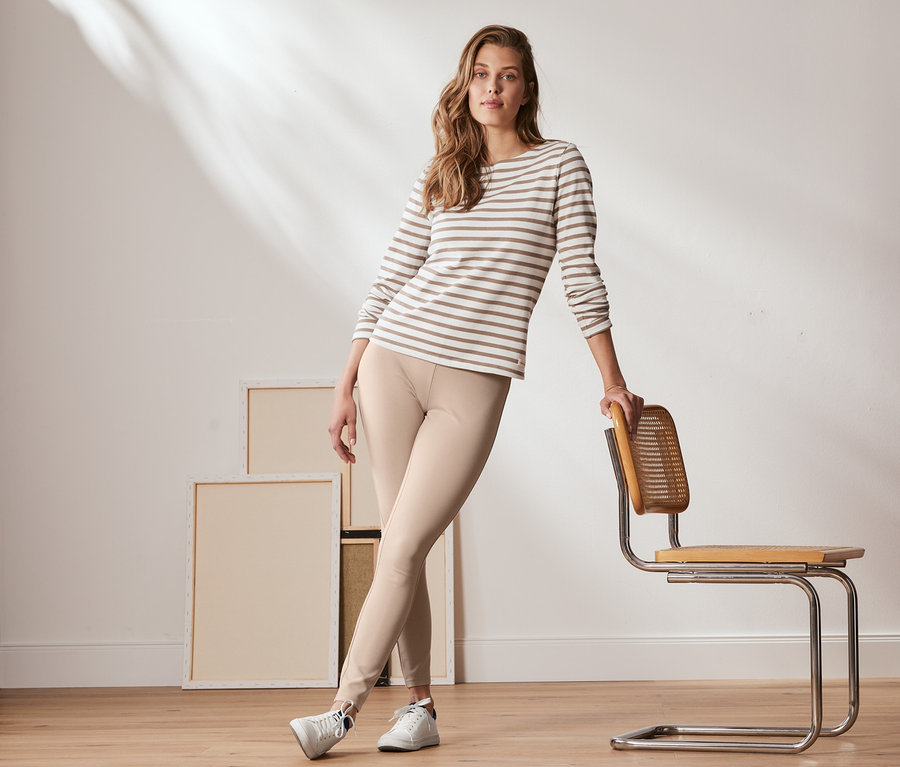 Frau posiert an einen Stuhl gelehnt in cremefarbene Basic-Leggings aus Heavy-Jersey und einem gestreiften Oberteil.