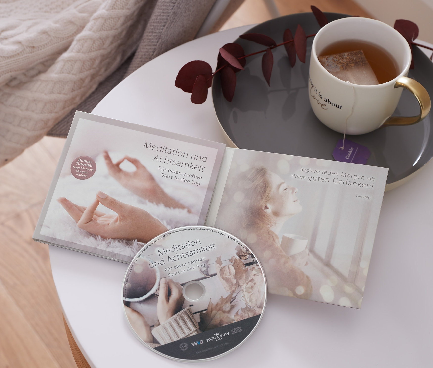 Auf einem weißen Tisch liegen die CD »Meditation und Achtsamkeit«, eine Tasse Tee und eine Strickdecke über einem Stuhl.