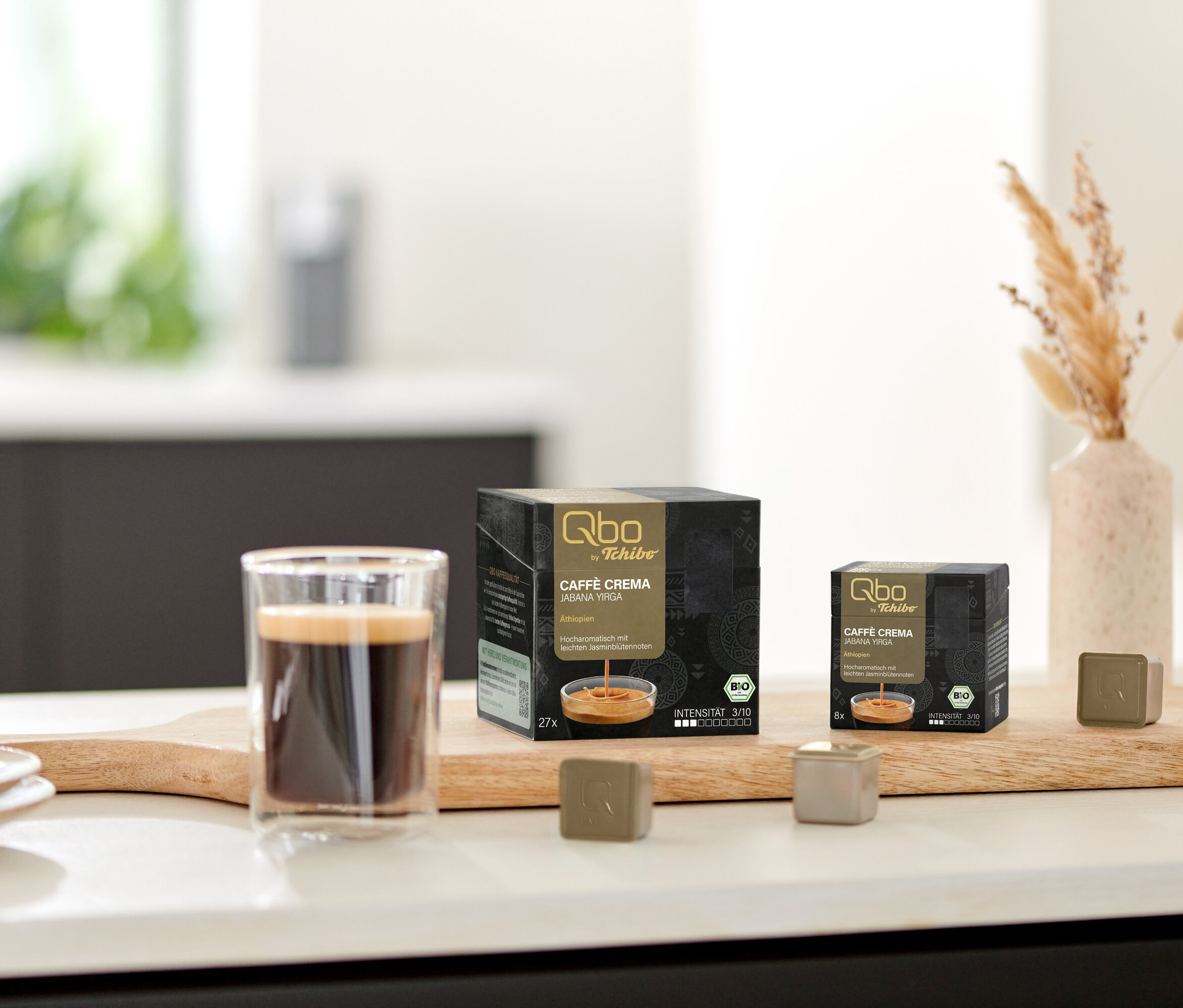 Auf einem Brett stehen eine Tasse Kaffee, eine Packung Qbo Caffè JABANA YIRGA - 27 Kapseln und eine Packung Qbo Caffè JABANA YIRGA - 8 Kapseln.