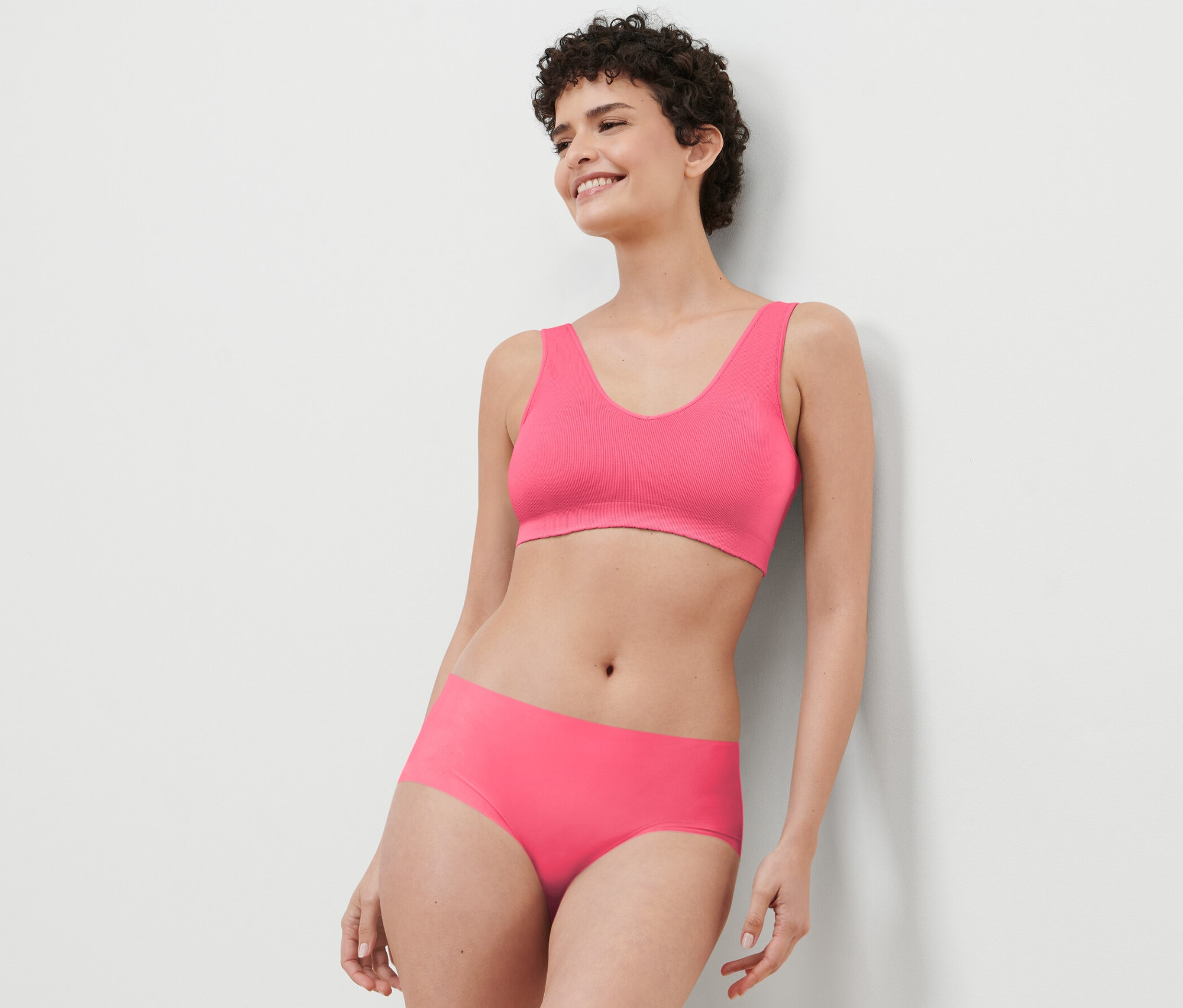 Frau in pinkem Sport-BH und 3 Pantys »Magic Cut« an eine weiße Wand gelehnt.