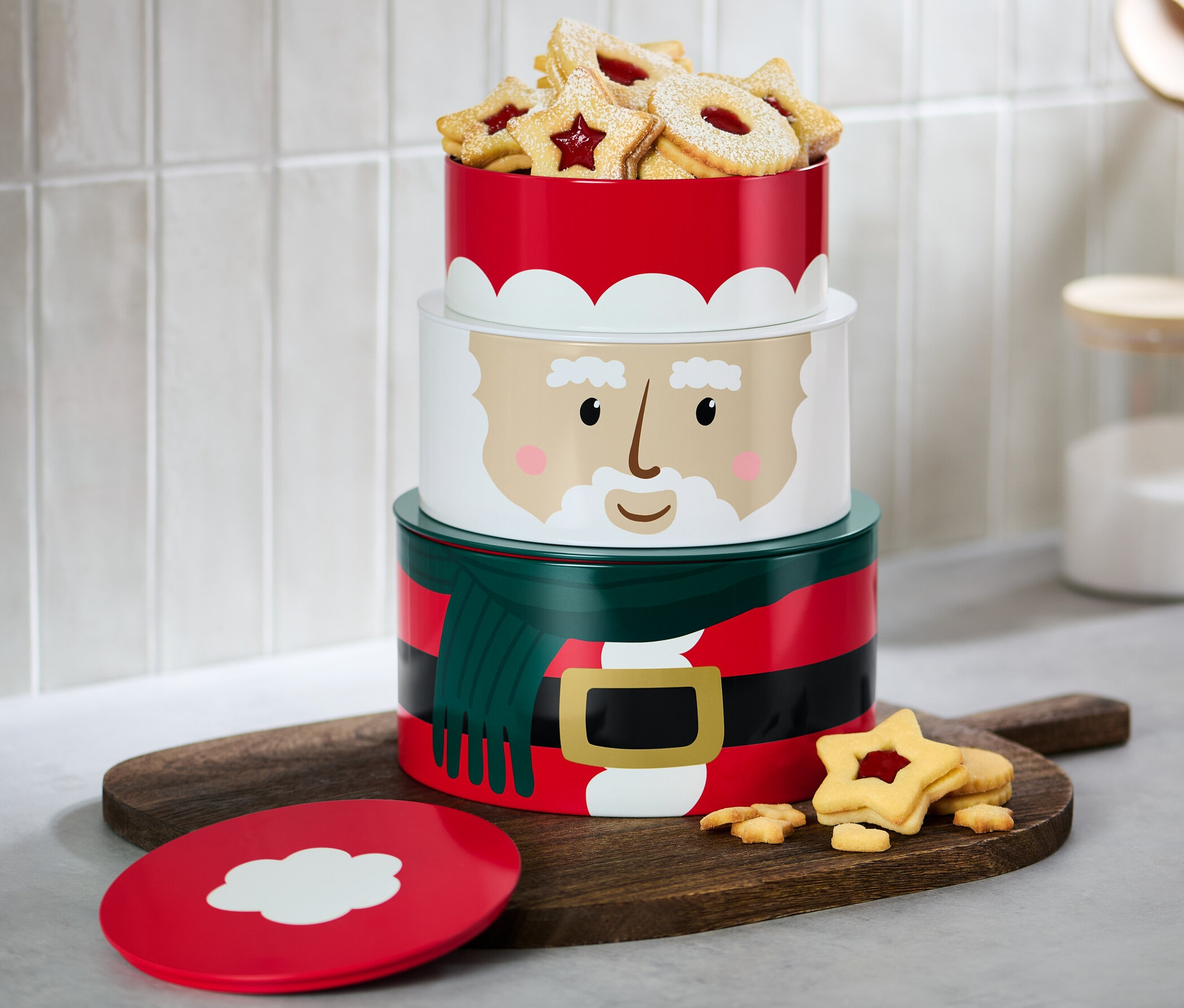 Dreistöckige Keksdosen mit Weihnachtsmotiv, gefüllt mit Keksen, stehen auf einem Holzbrett.