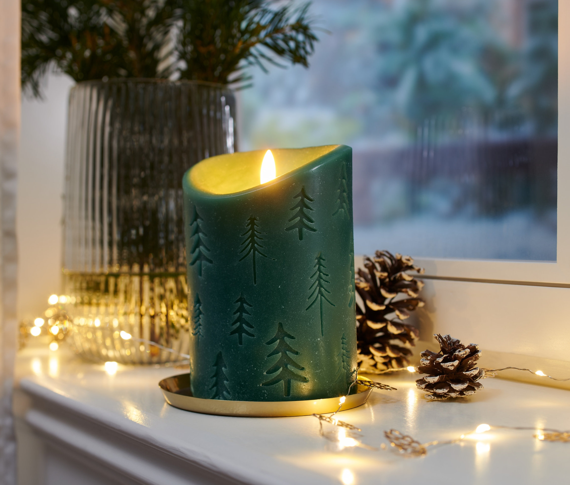 Eine grüne LED-Echtwachskerze mit Tannenbaum Muster steht auf einem goldenen Teller, neben Tannenzapfen und Lichtern.