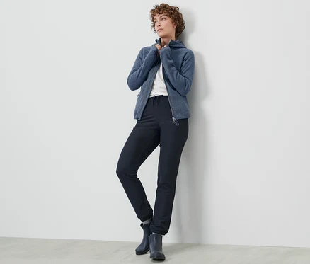 Frau posiert in Softshell-Joggpants und blauer Reißverschlussjacke an einer Wand.