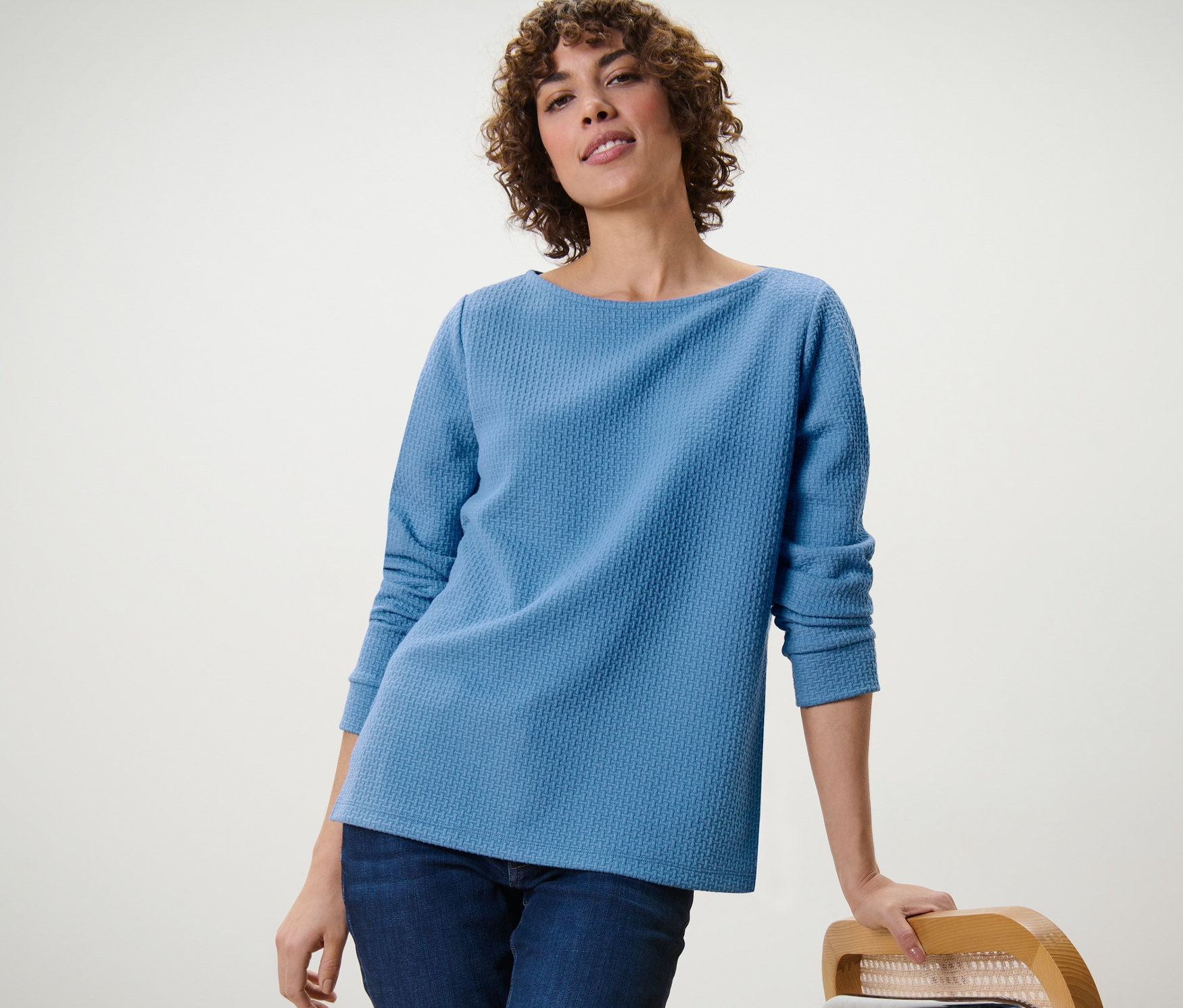 Eine Frau, die sich an einen Stuhl lehnt, trägt ein blaues, strukturiertes Sweatshirt.