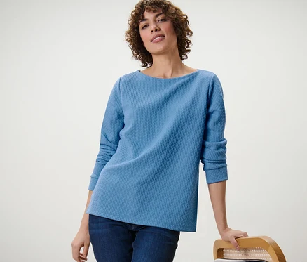 Eine Frau, die sich an einen Stuhl lehnt, trägt ein blaues, strukturiertes Sweatshirt.