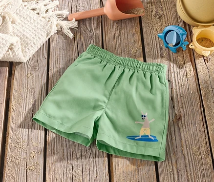 Grüne Kinder-Boardshorts mit Känguru-Motiv auf Surfbrett liegen auf einem Holztisch mit Sandspielzeug.