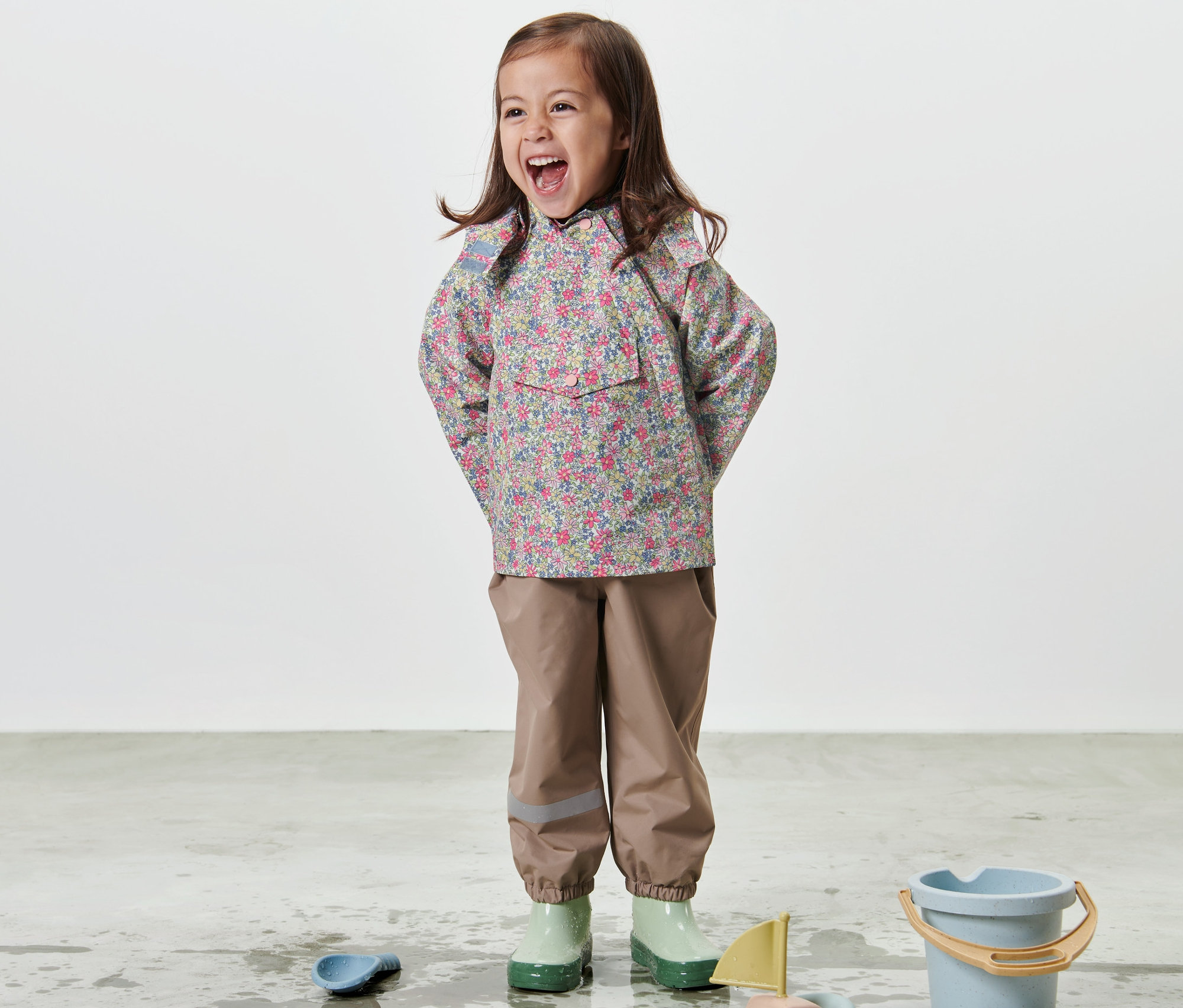 Lachendes Mädchen steht in Kinder-Regenjacke mit Blumenmuster und spielt mit Spielzeug auf dem Boden.