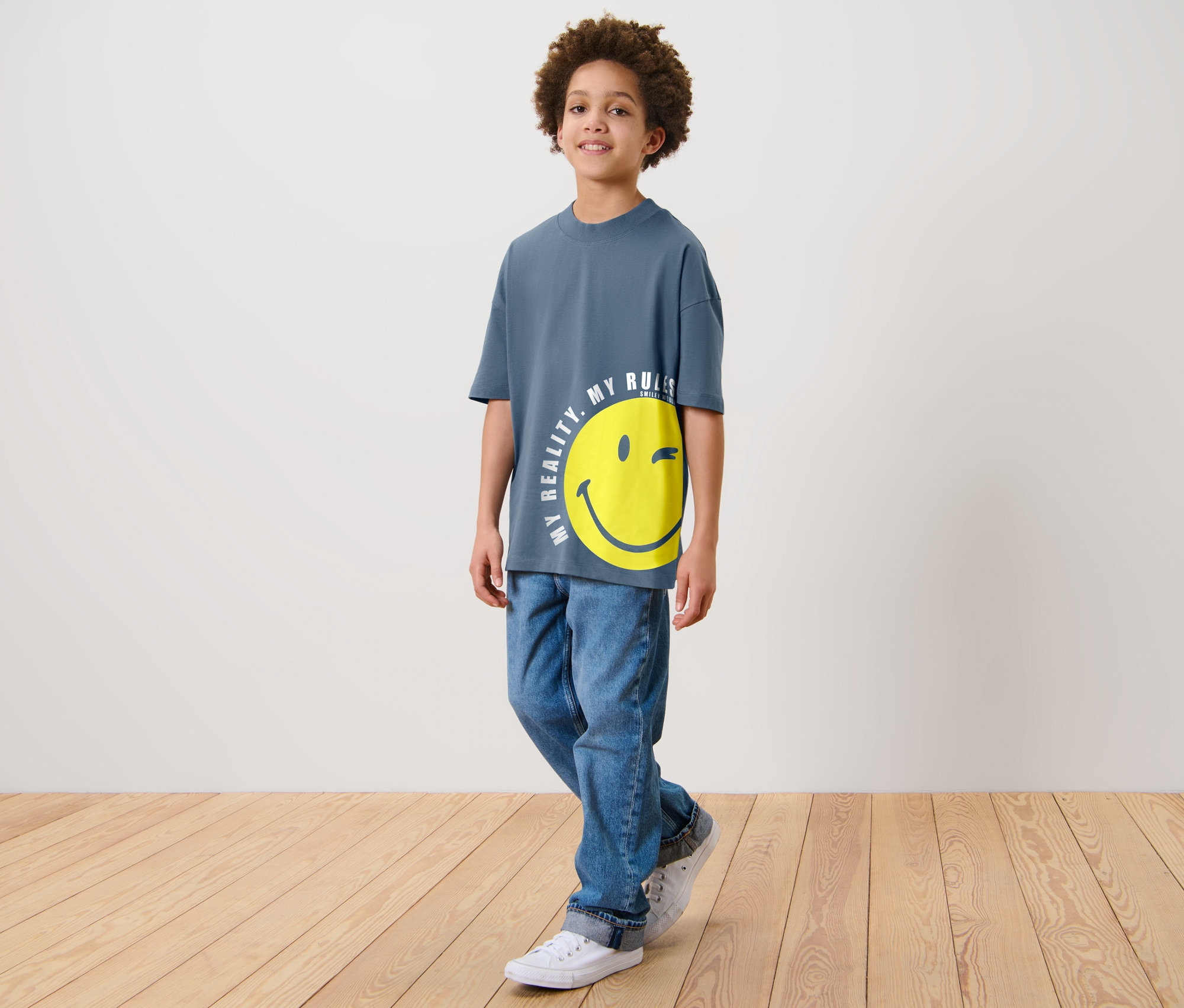 Ein Junge steht und trägt ein blaues T-Shirt mit einem gelben Smiley-Aufdruck und Jeans. Er trägt 2 Kinder-T-Shirts »Smiley«.