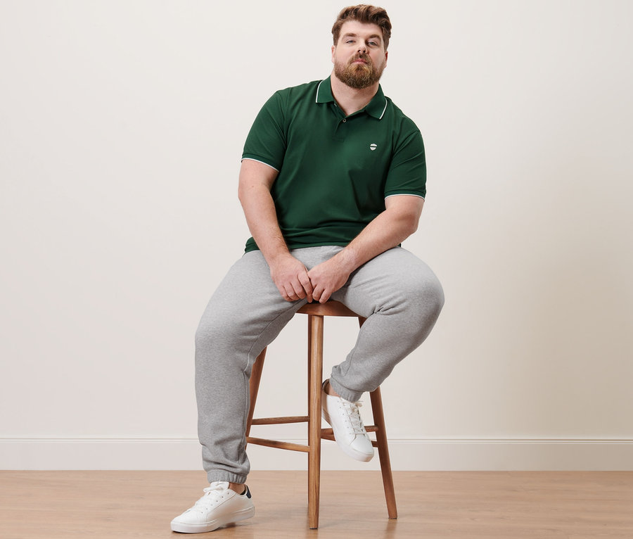 Mann sitzt auf einem Hocker und trägt ein dunkelgrünes Piqué-Poloshirt und eine Sweathose.